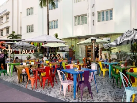 Outdoor restaurant with colorful metal chairs and tables, striped umbrellas, and plants on patterned tile flooring, set against a white art-deco building with large windows. People are dining and walking nearby.
