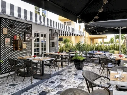 Outdoor cafe with patterned tile floor, black-and-white striped awning, tables and chairs, potted plants, and decorated wall with framed pictures. Sunlight brightens the area, giving it a cozy, inviting atmosphere.